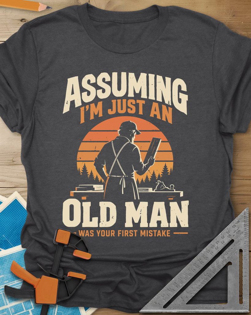 Old Man Mistake t-shirt in Dark Heather Grey, a woodworker in an apron holding a board before a striped sunset