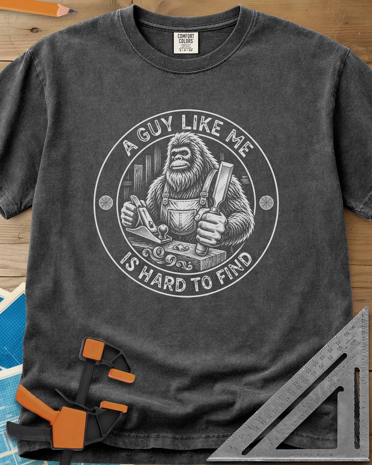 A Guy Like Me Is Hard To Find Woodworking heavyweight tee in Pepper, Bigfoot in overalls with a hand plane and chisel inside