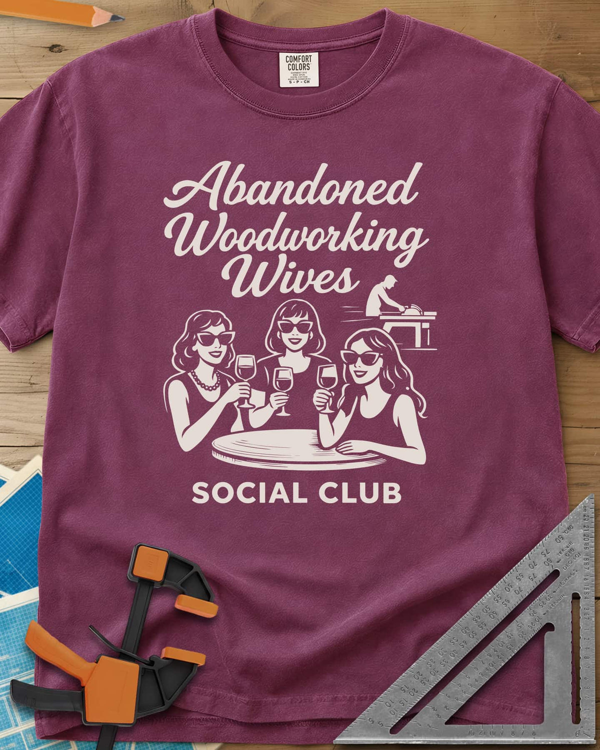 Abandoned Woodworking Wives heavyweight tee in Berry, three women in sunglasses with wine, a man at a table saw behind