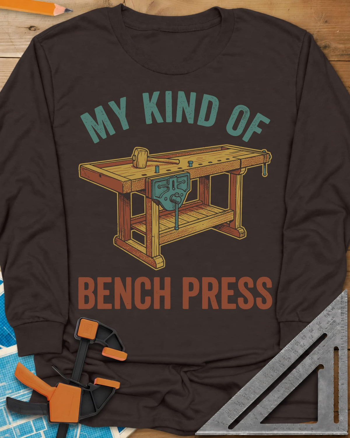 Bench Press long sleeve shirt in Dark Chocolate, a wooden workbench with a vise and mallet between the two lines