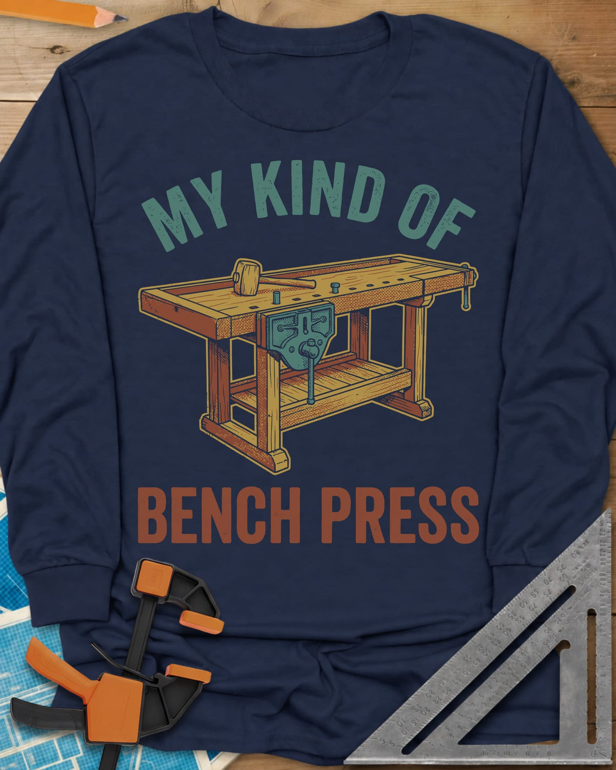 Bench Press long sleeve shirt in Navy, a wooden workbench with a vise and mallet between the two lines