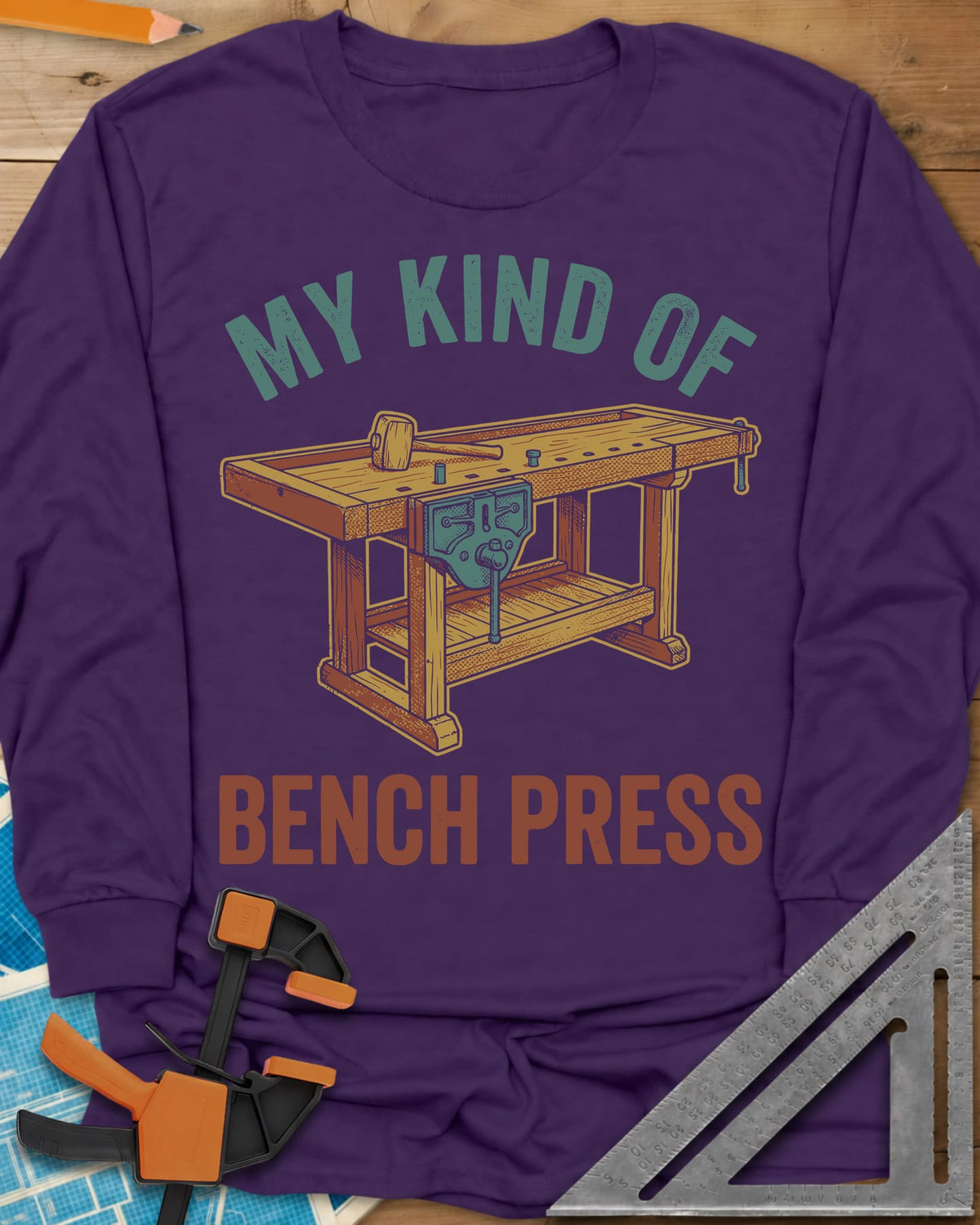 Bench Press long sleeve shirt in Purple, a wooden workbench with a vise and mallet between the two lines