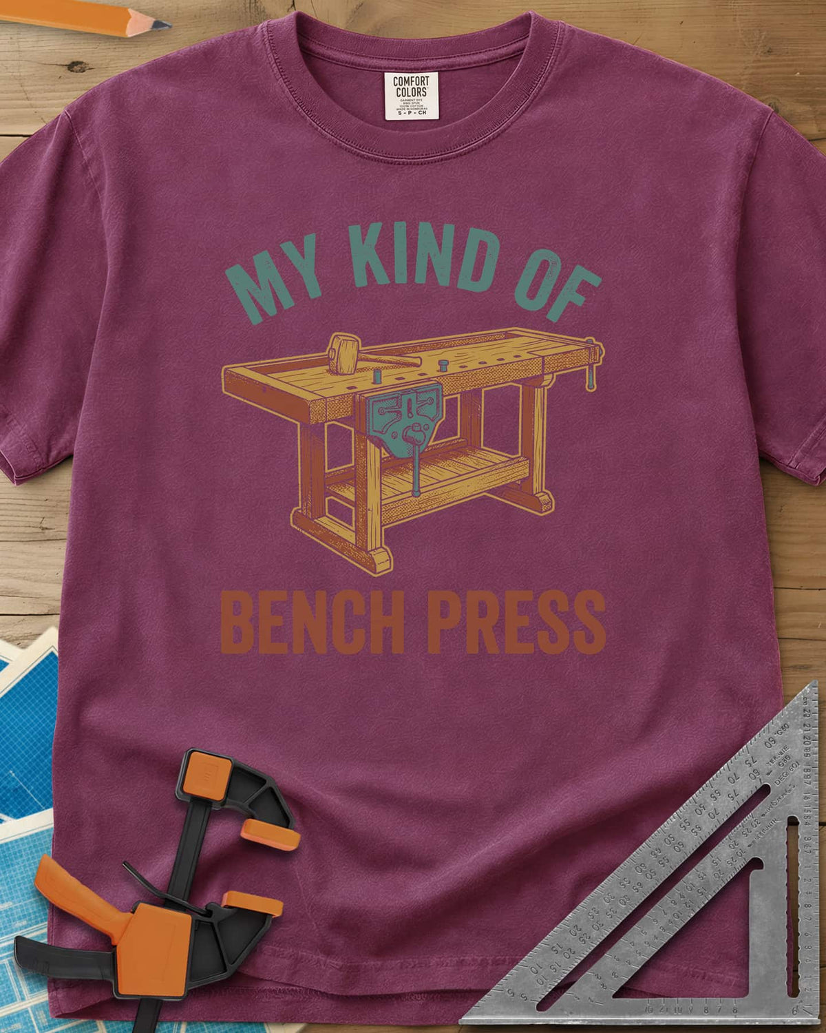 Bench Press heavyweight tee in Berry, a wooden workbench with a vise and mallet between the two lines