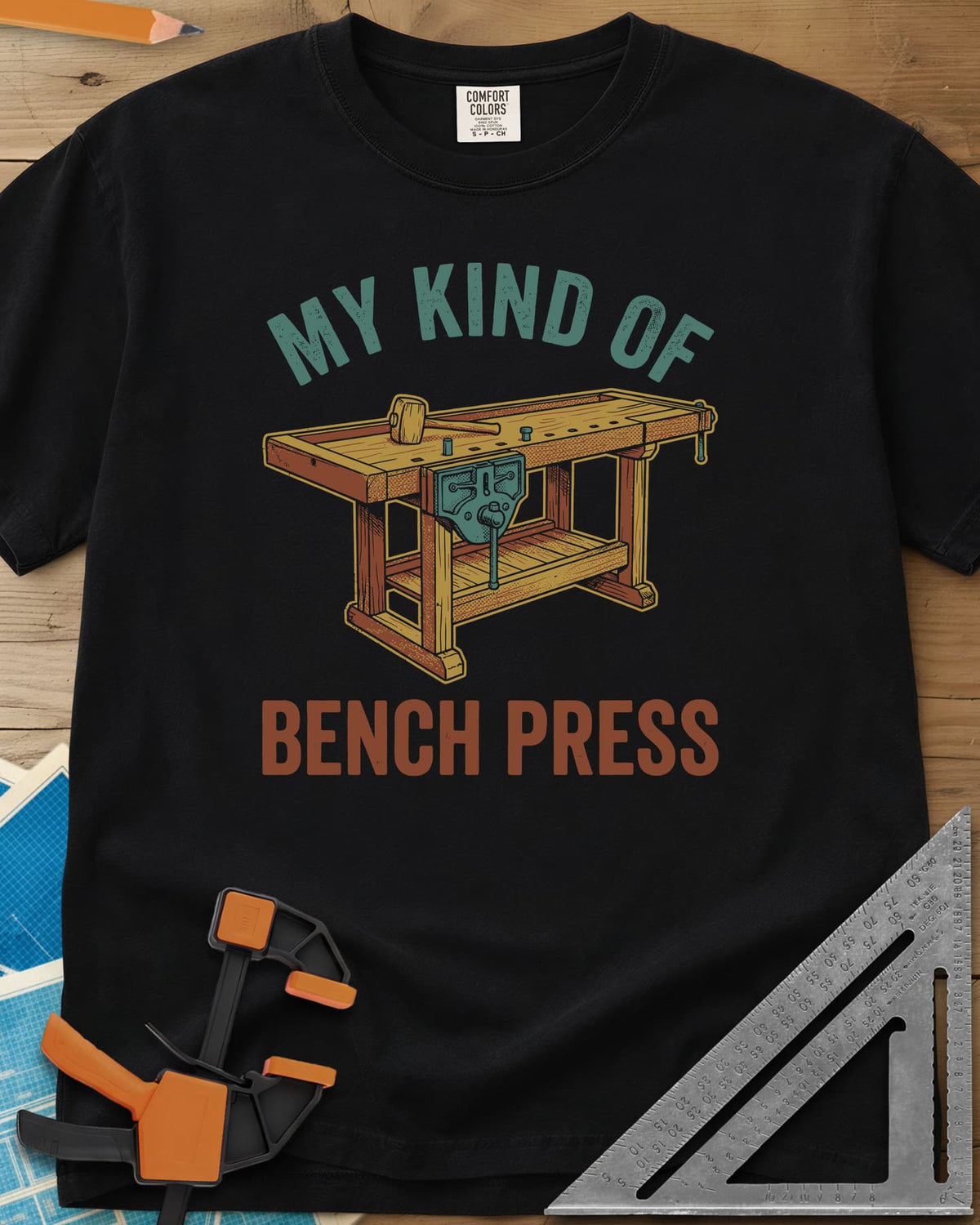 Bench Press heavyweight tee in Black, a wooden workbench with a vise and mallet between the two lines