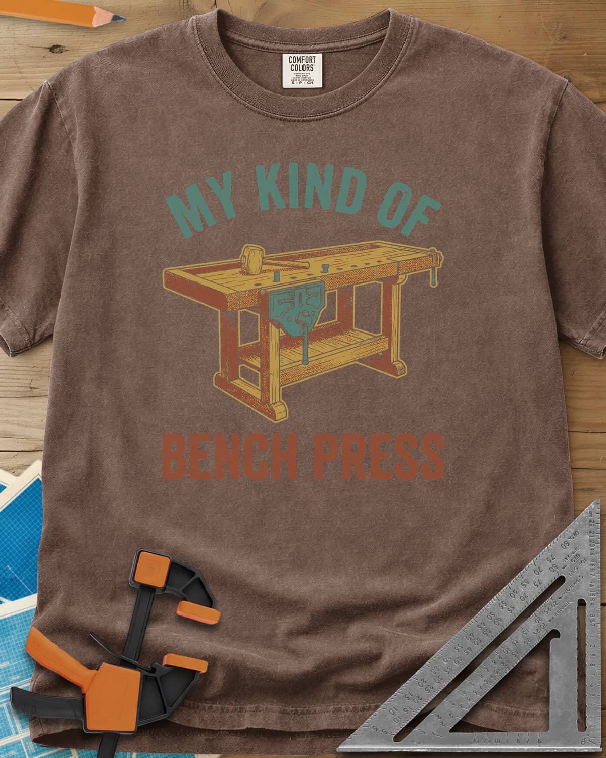 Bench Press heavyweight tee in Espresso, a wooden workbench with a vise and mallet between the two lines