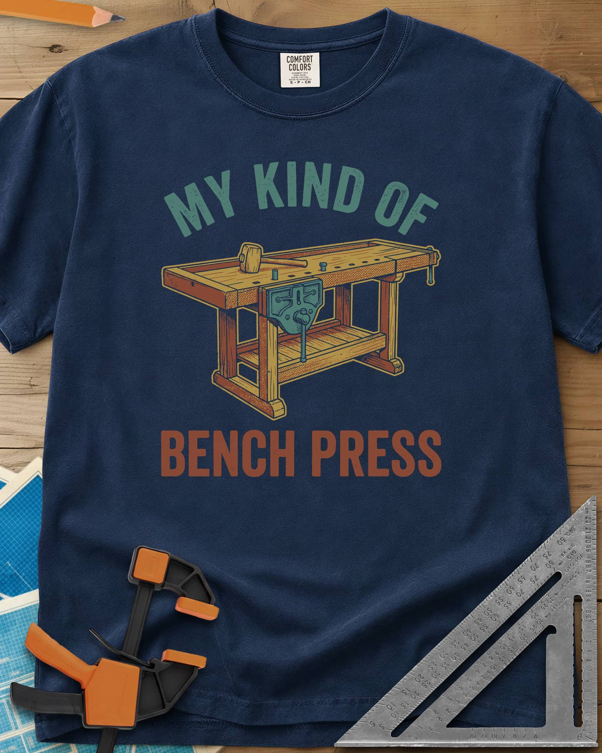 Bench Press heavyweight tee in Navy, a wooden workbench with a vise and mallet between the two lines