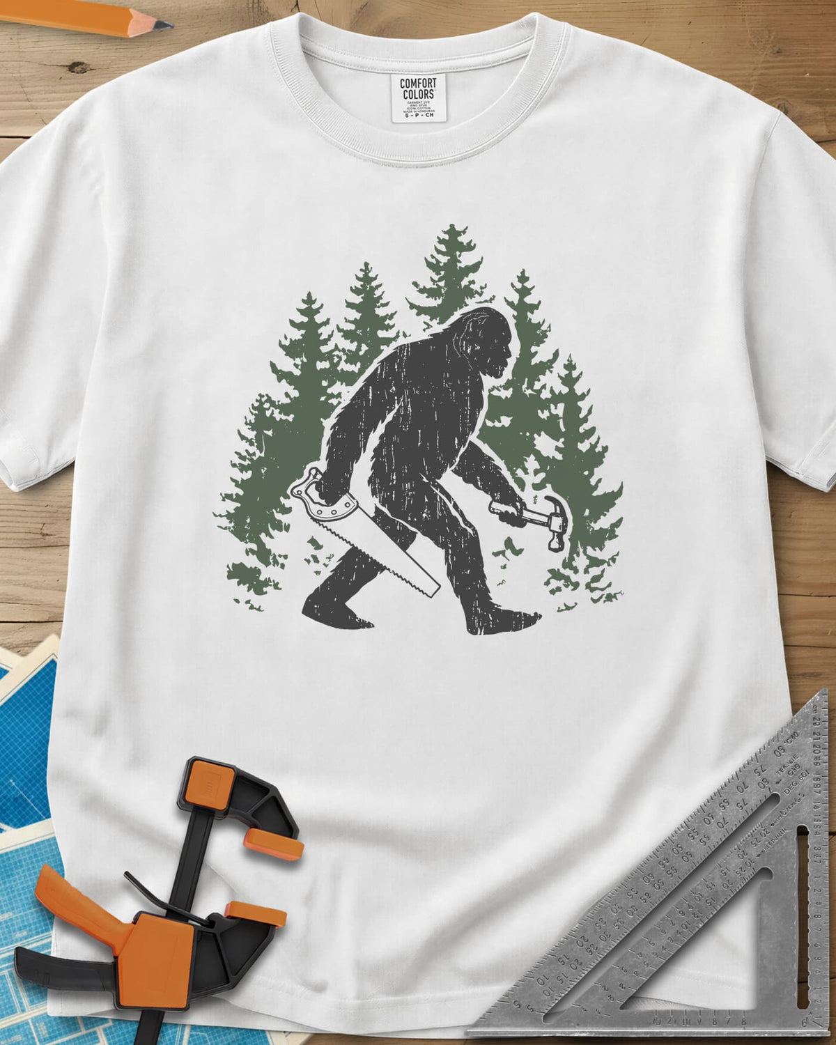 Big Foot heavyweight tee in White, Bigfoot walking through pine trees with a hand saw and a hammer