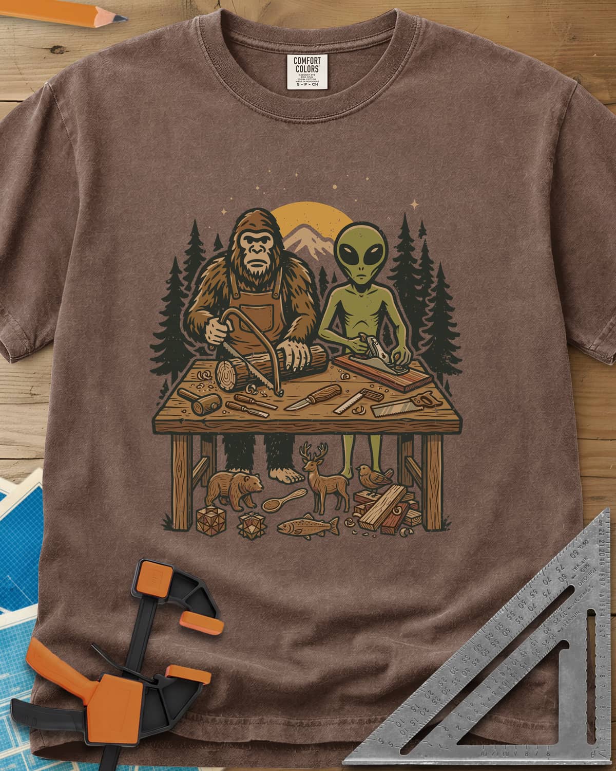 Bigfoot And Alien Woodshop heavyweight tee in Espresso, Bigfoot sawing a log and an alien planing a board at one bench