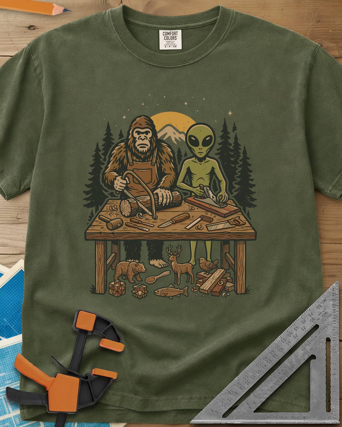 Bigfoot And Alien Woodshop heavyweight tee in Hemp, Bigfoot sawing a log and an alien planing a board at one bench