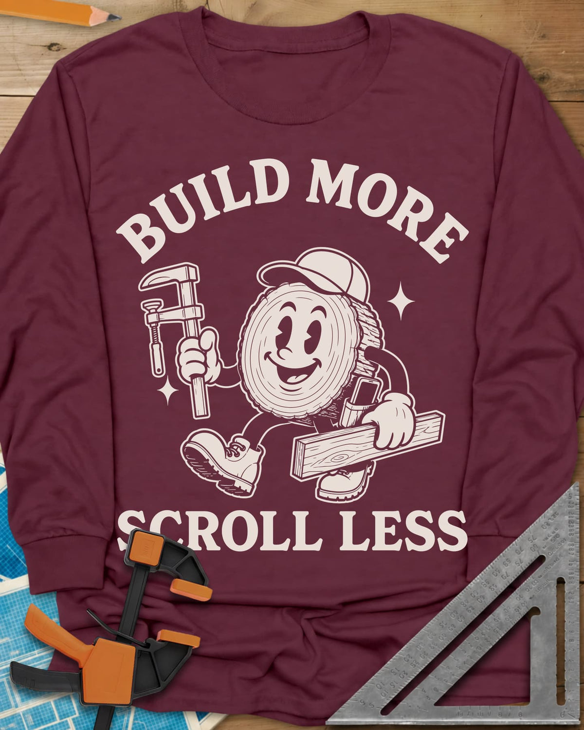Build More Scroll Less long sleeve shirt in Maroon, a smiling log-slice mascot in boots carrying a tool and a board