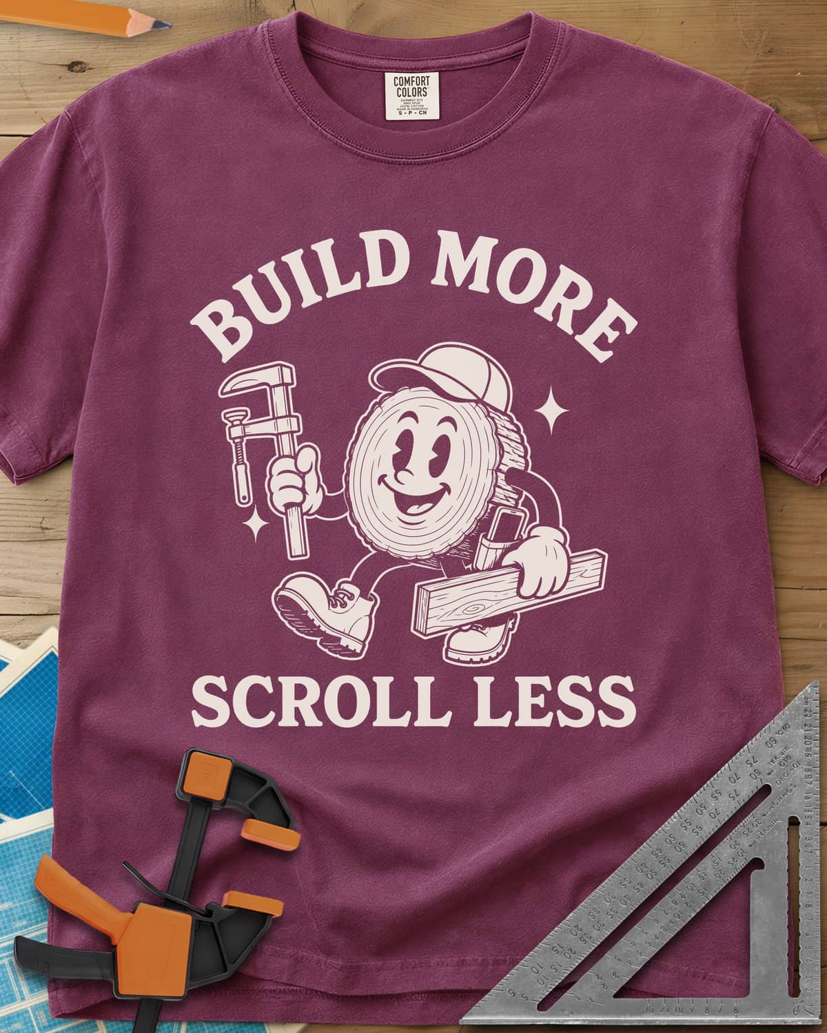 Build More Scroll Less heavyweight tee in Berry, a smiling log-slice mascot in boots carrying a tool and a board