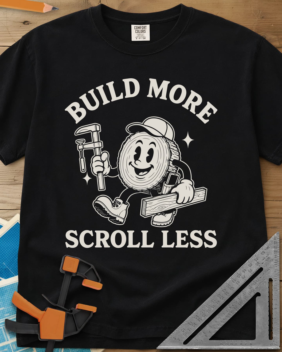 Build More Scroll Less heavyweight tee in Black, a smiling log-slice mascot in boots carrying a tool and a board