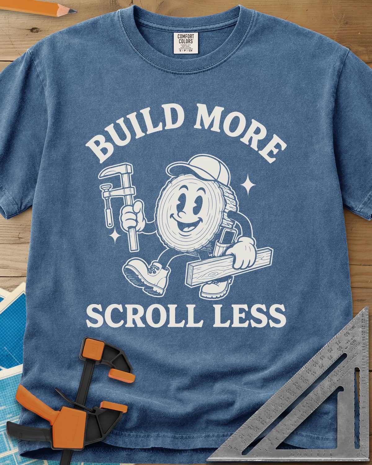 Build More Scroll Less heavyweight tee in Blue Jean, a smiling log-slice mascot in boots carrying a tool and a board