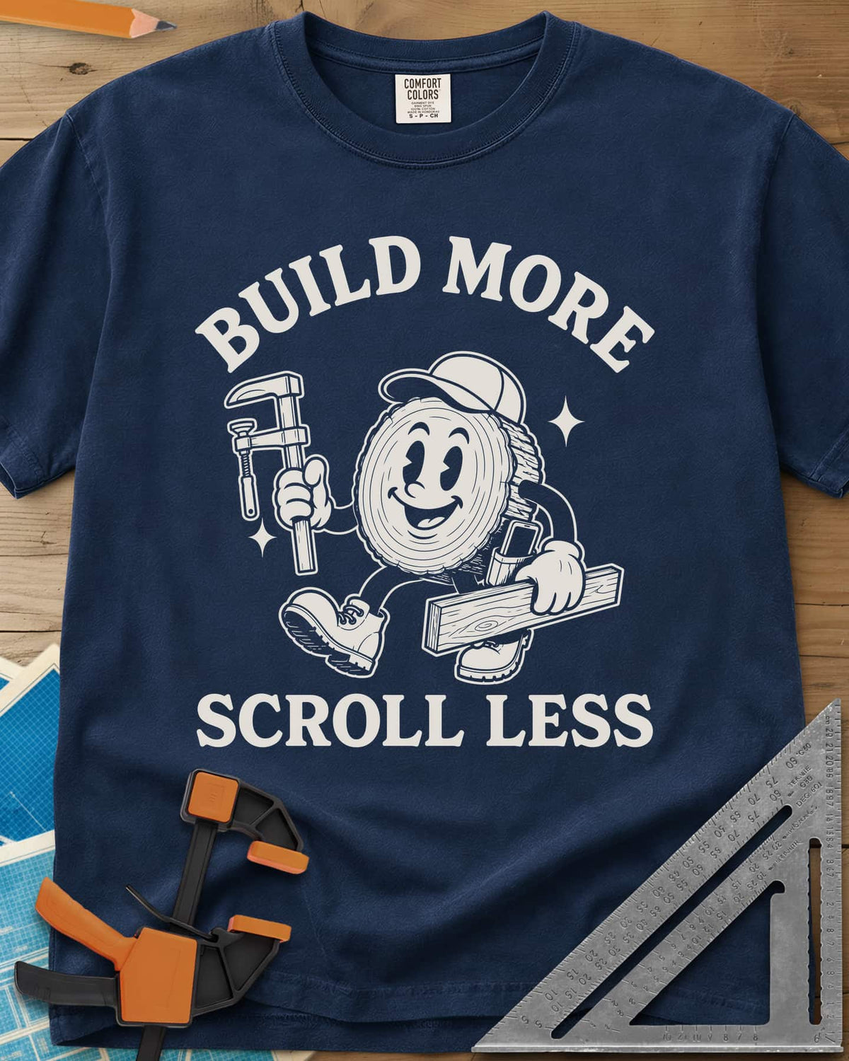 Build More Scroll Less heavyweight tee in Navy, a smiling log-slice mascot in boots carrying a tool and a board