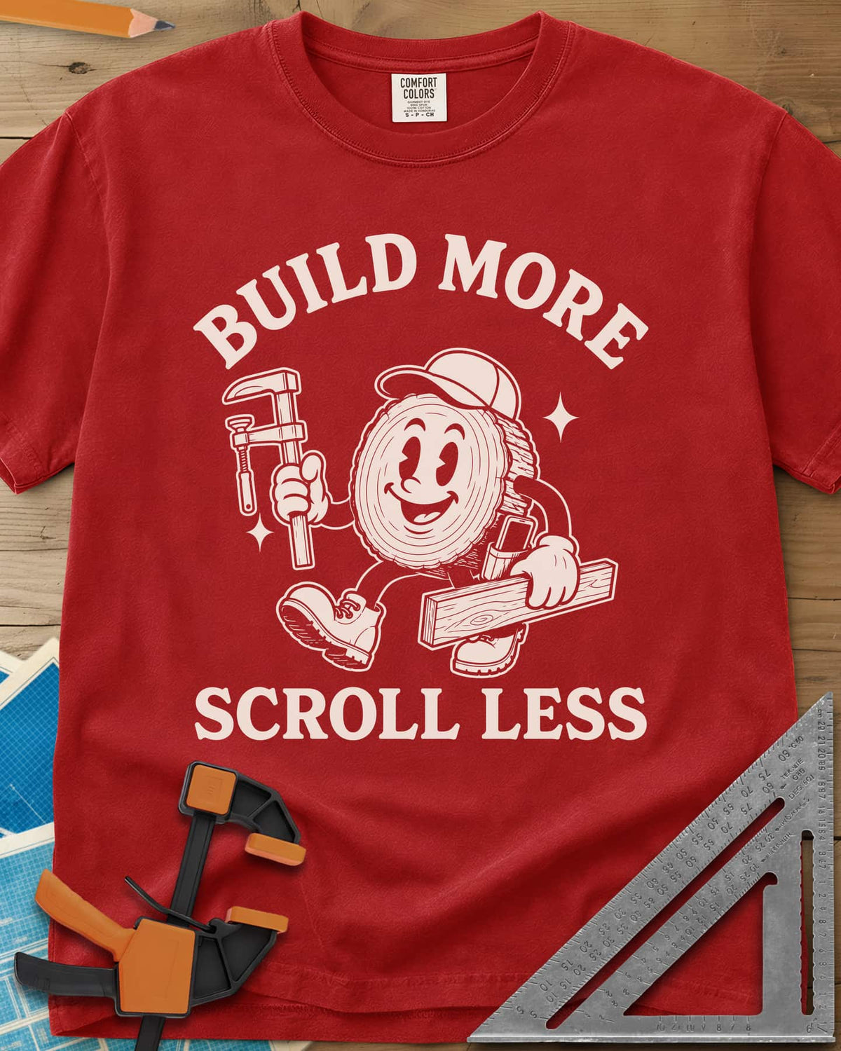 Build More Scroll Less heavyweight tee in Red, a smiling log-slice mascot in boots carrying a tool and a board
