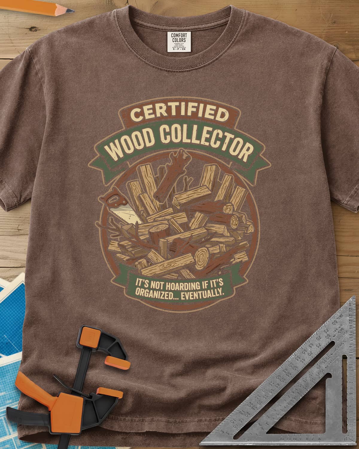 Certified Wood Collector heavyweight tee in Espresso, ribbon banners over a heap of logs, offcuts and a handsaw