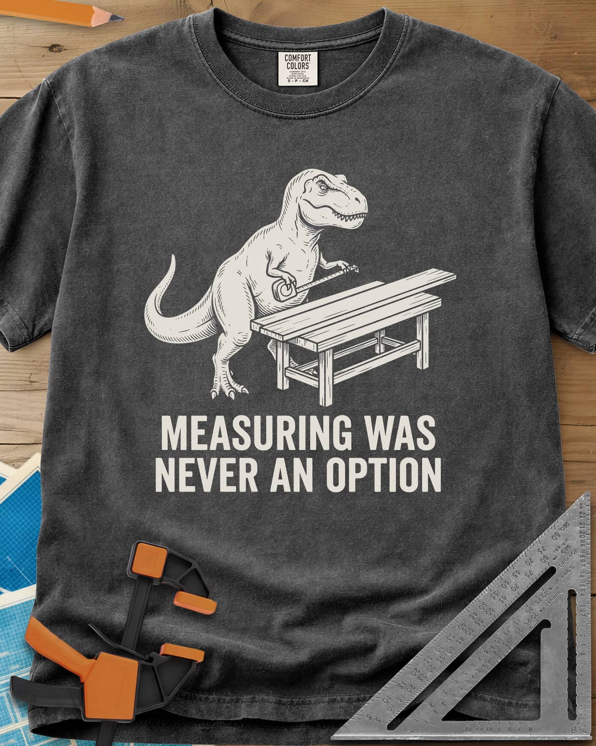 Dino Measuring heavyweight tee in Pepper, a T. rex with a tape measure beside a plank workbench