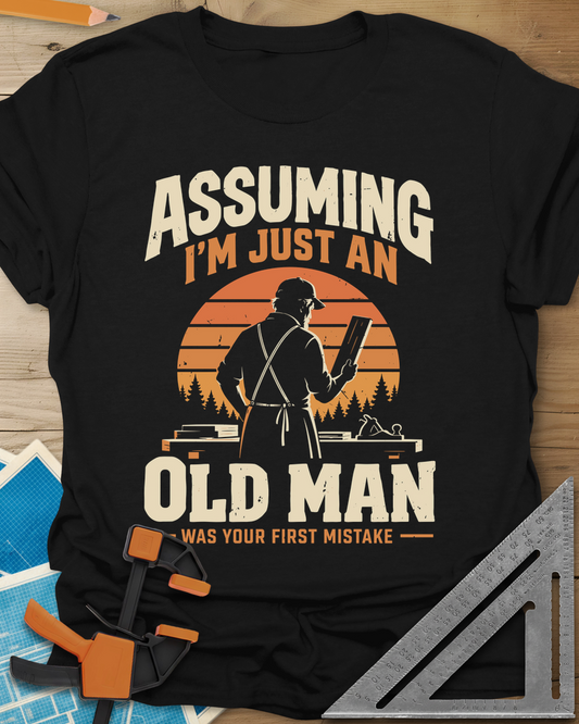 Old Man Mistake t-shirt in Black, a woodworker in an apron holding a board before a striped sunset