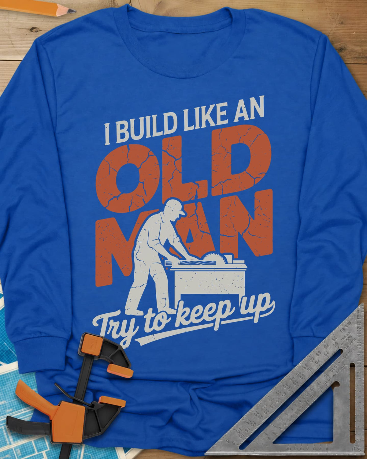 I Build Like An Old Man Woodworking long sleeve shirt in Royal, a man in a cap pushing a board through a table saw