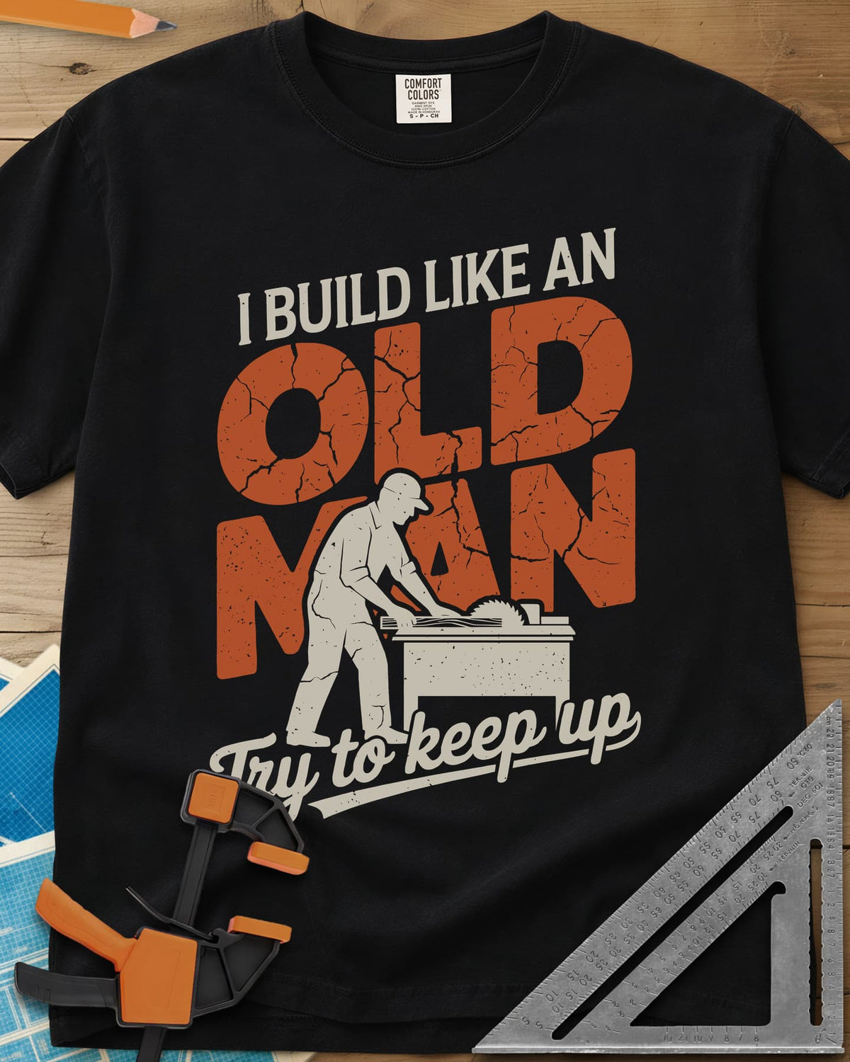 I Build Like An Old Man Woodworking heavyweight tee in Black, a man in a cap pushing a board through a table saw