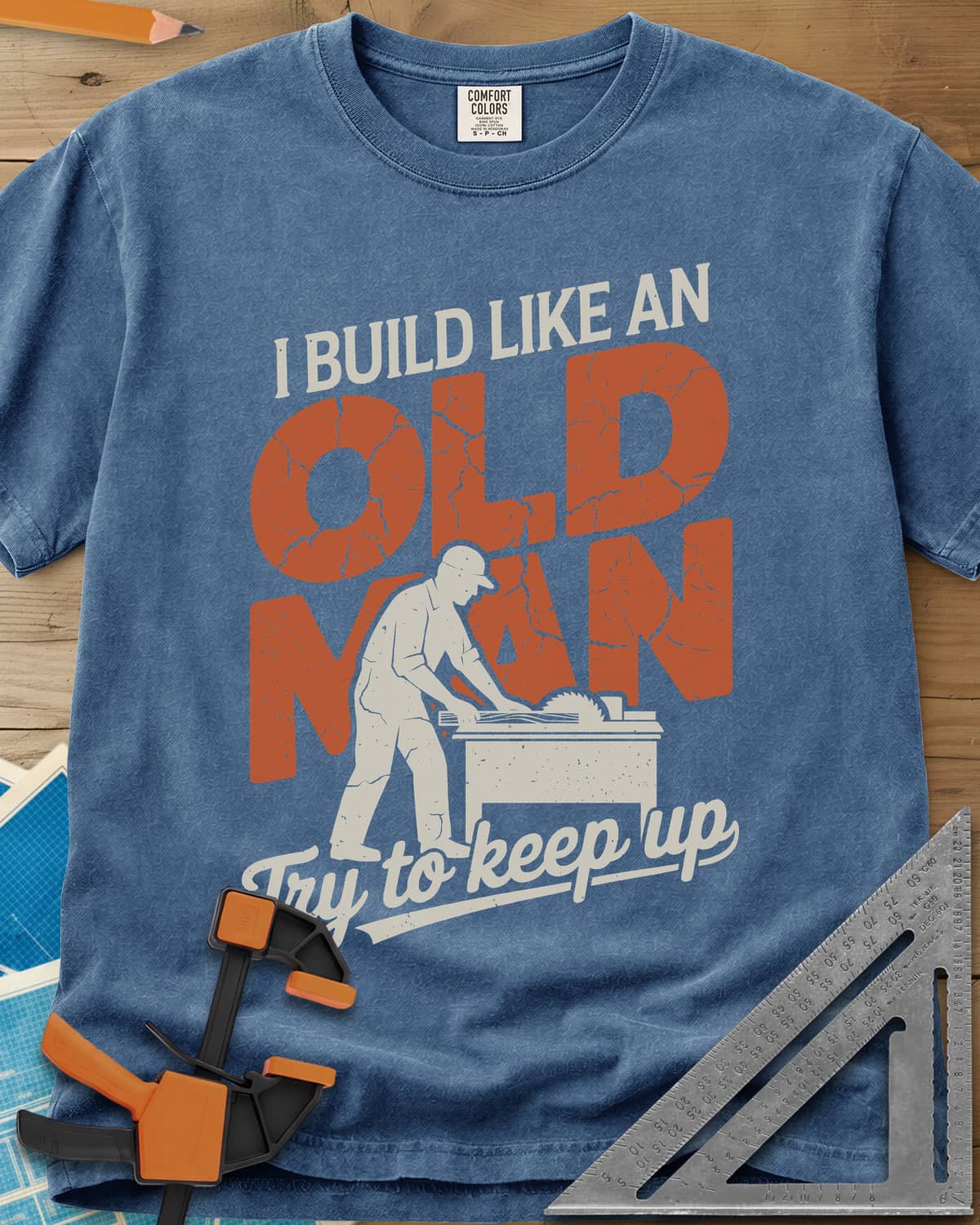 I Build Like An Old Man Woodworking heavyweight tee in Blue Jean, a man in a cap pushing a board through a table saw