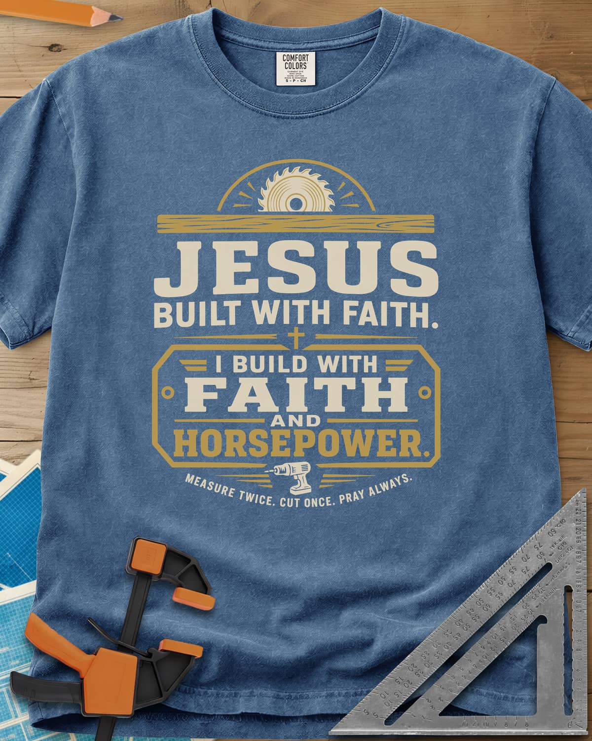 I Build With Faith And Horsepower heavyweight tee in Blue Jean, saw blade over a plank above a plaque with a cordless drill