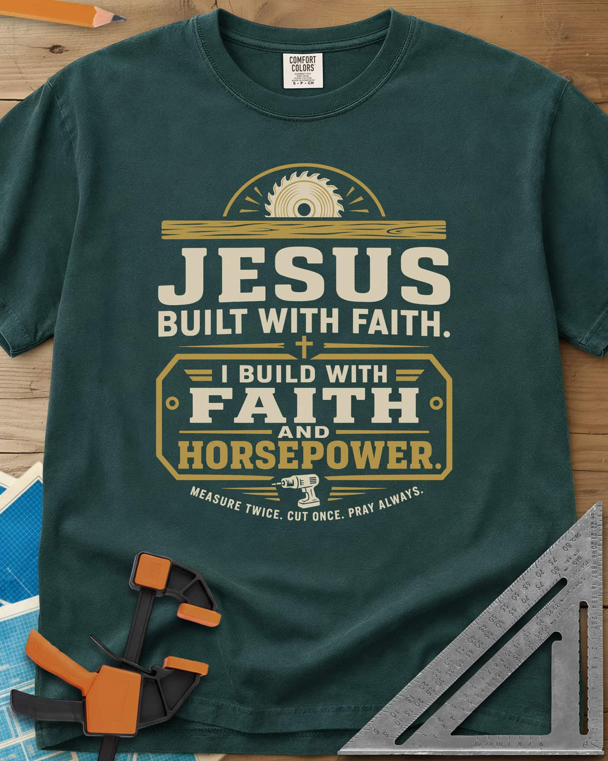 I Build With Faith And Horsepower heavyweight tee in Blue Spruce, saw blade over a plank above a plaque with a cordless drill
