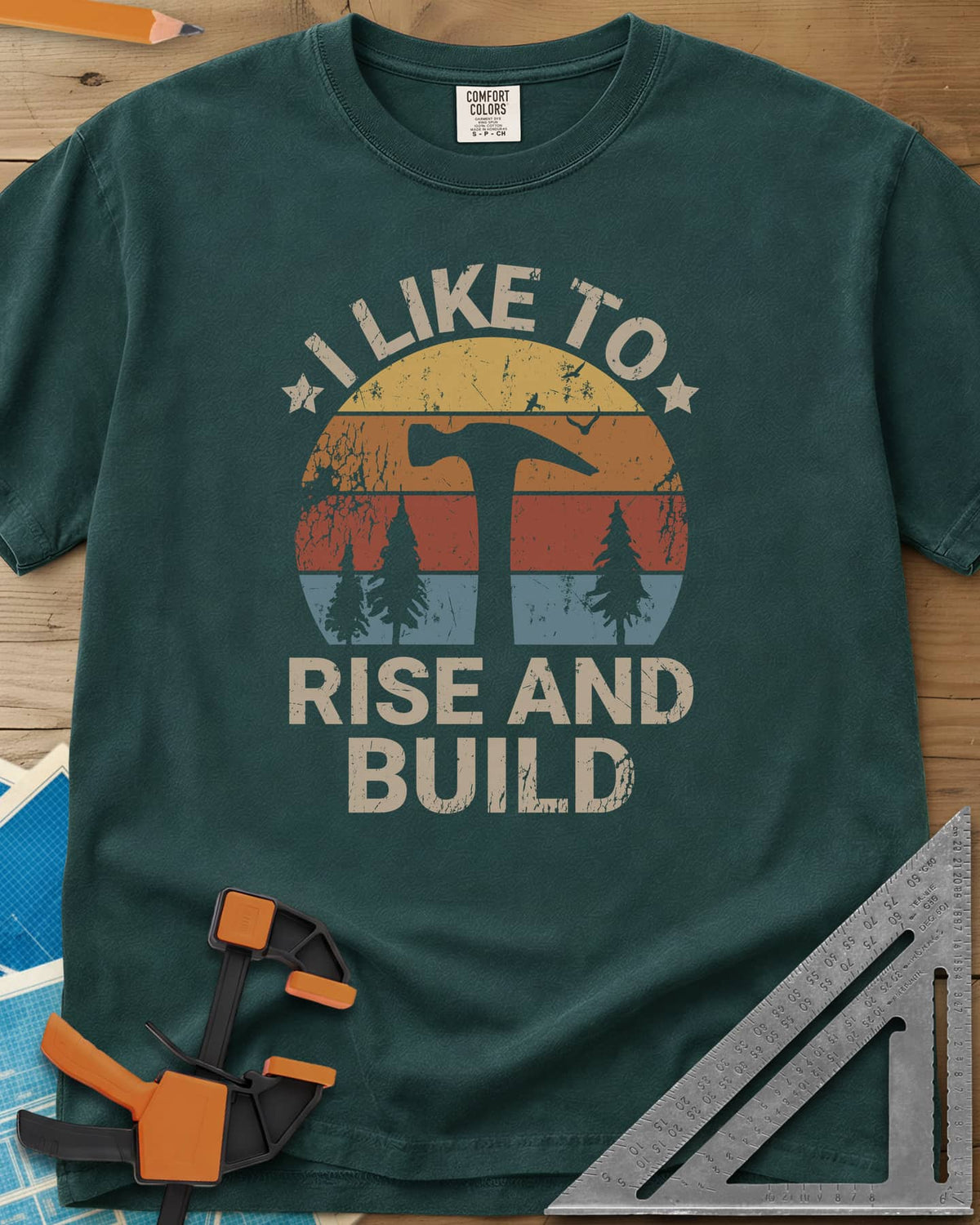 I Like To Rise And Build heavyweight tee in Blue Spruce, a retro striped sunset with a hammer silhouette and pine trees