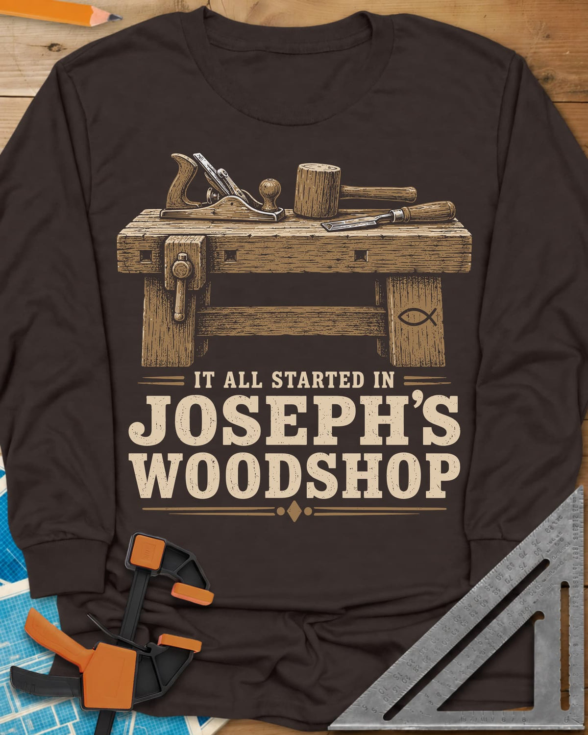 Joseph's Woodshop long sleeve shirt in Dark Chocolate, a workbench with plane, mallet and chisel and a small fish symbol