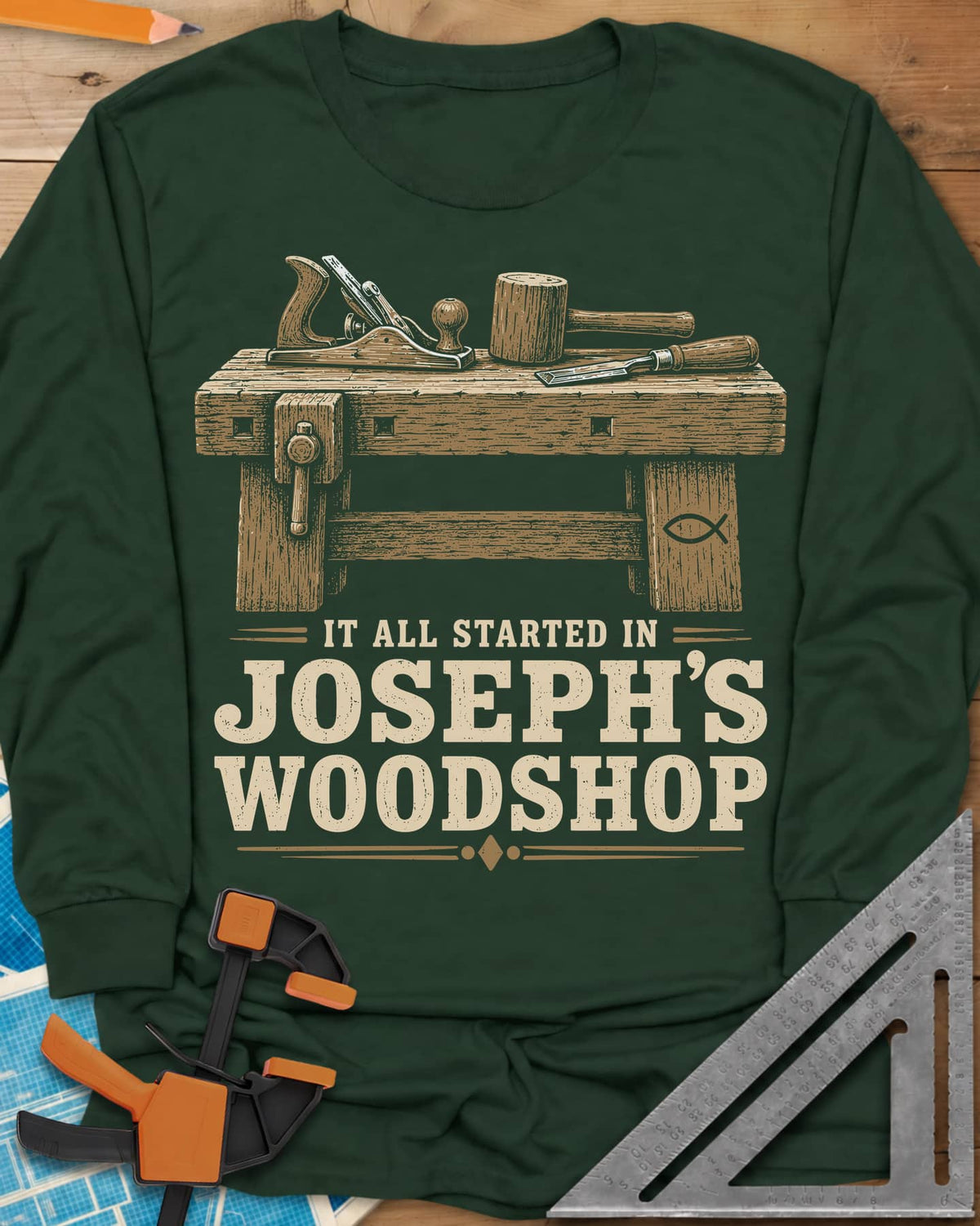 Joseph's Woodshop long sleeve shirt in Forest Green, a workbench with plane, mallet and chisel and a small fish symbol