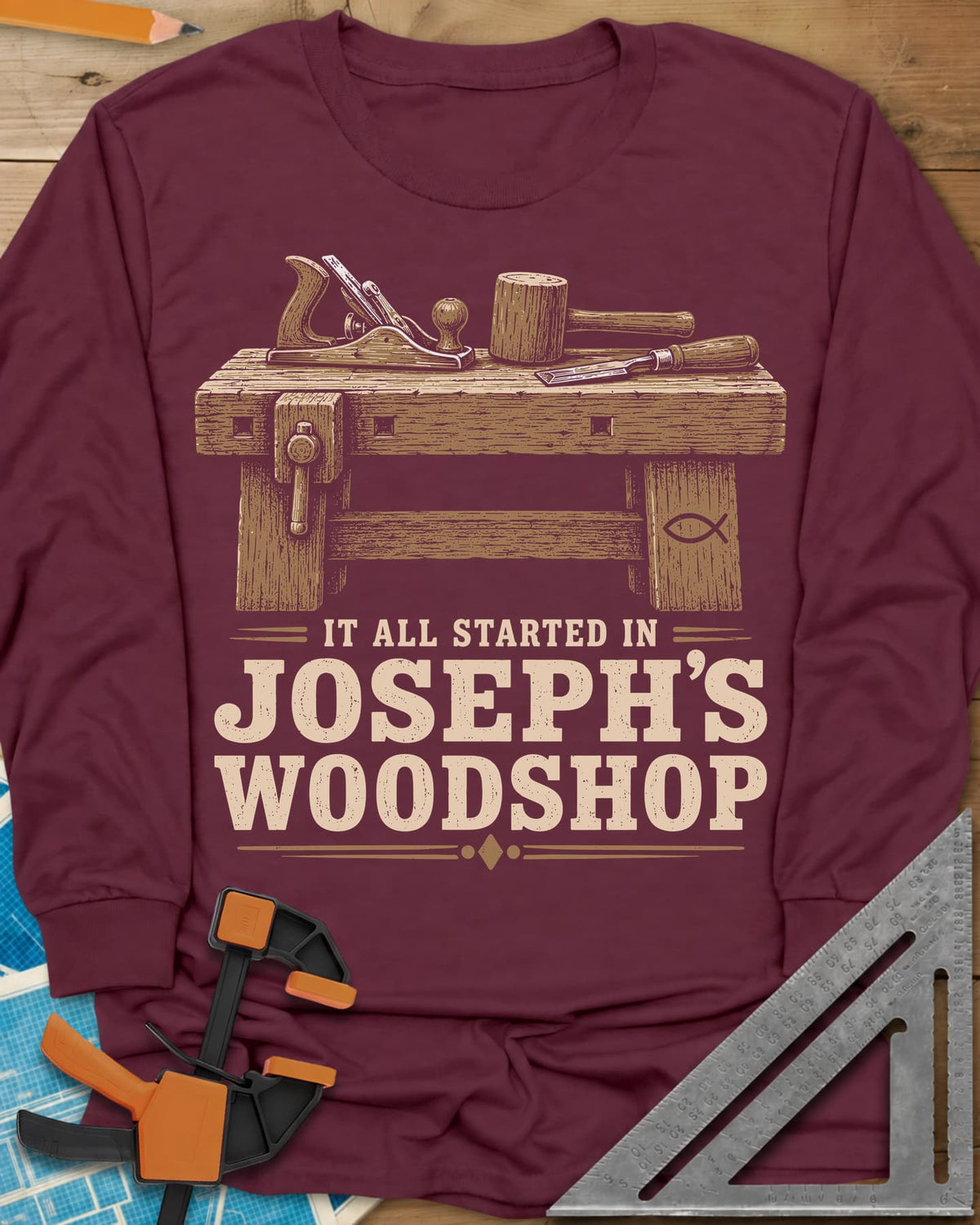 Joseph's Woodshop long sleeve shirt in Maroon, a workbench with plane, mallet and chisel and a small fish symbol