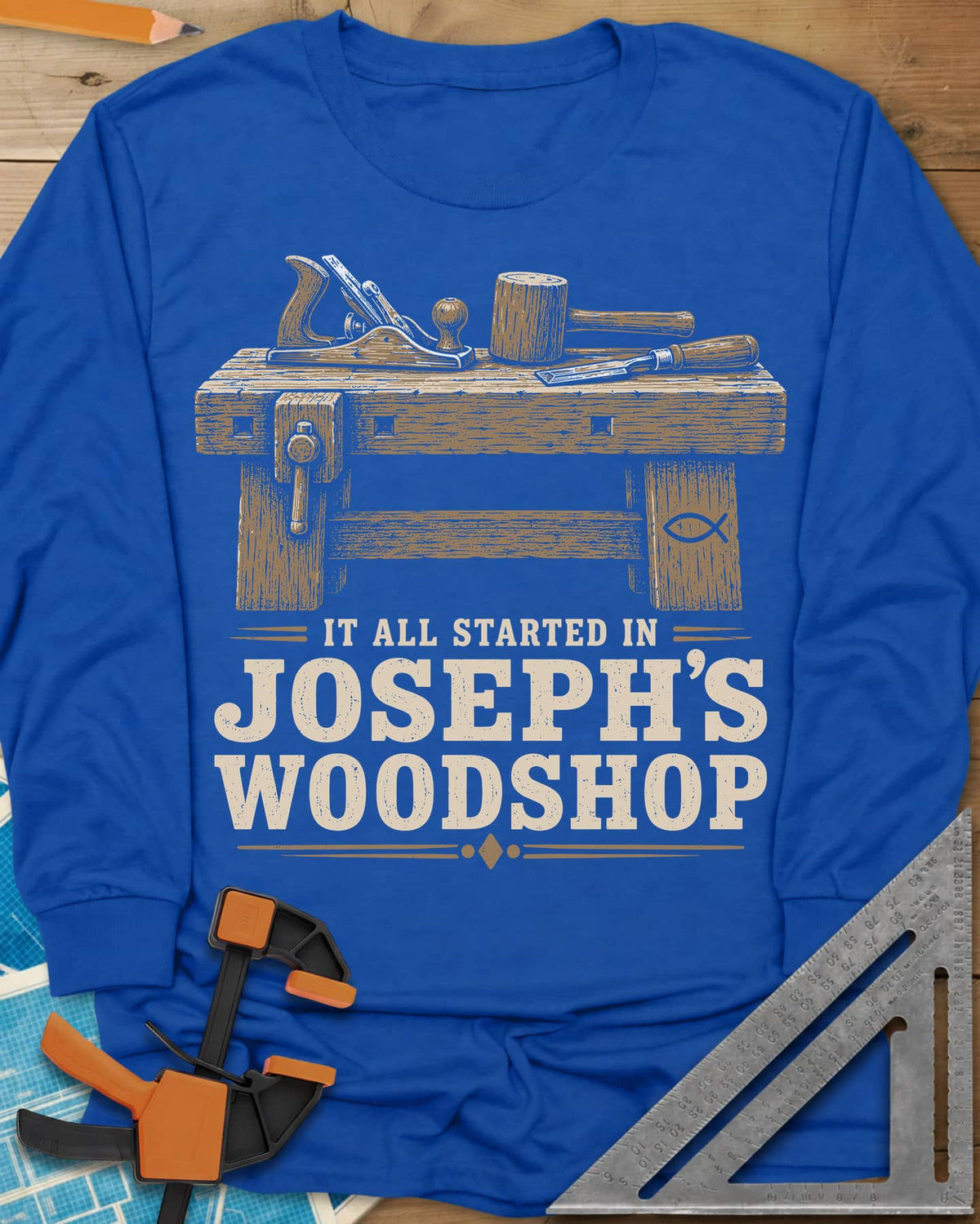 Joseph's Woodshop long sleeve shirt in Royal, a workbench with plane, mallet and chisel and a small fish symbol