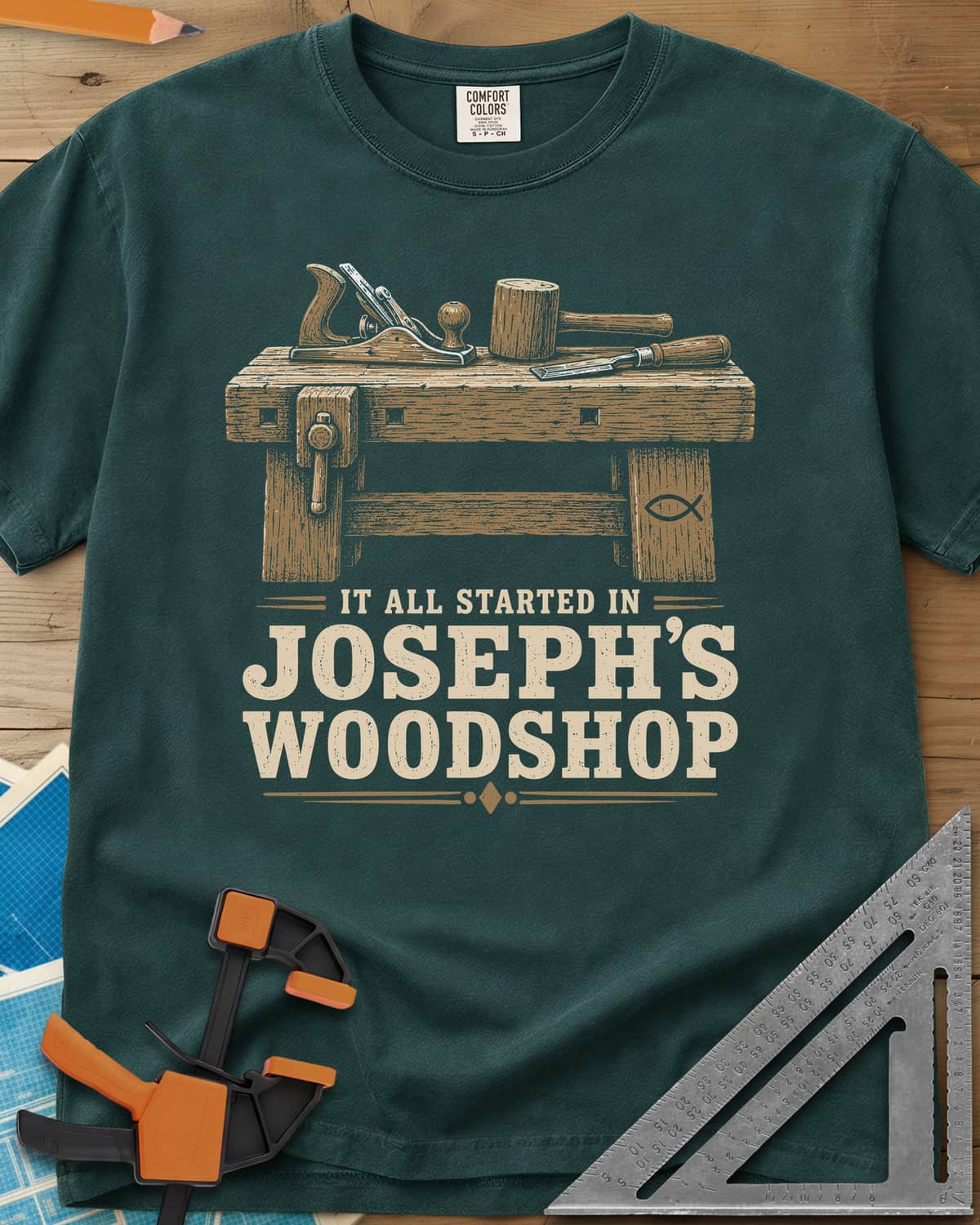 Joseph's Woodshop heavyweight tee in Blue Spruce, a workbench with plane, mallet and chisel and a small fish symbol