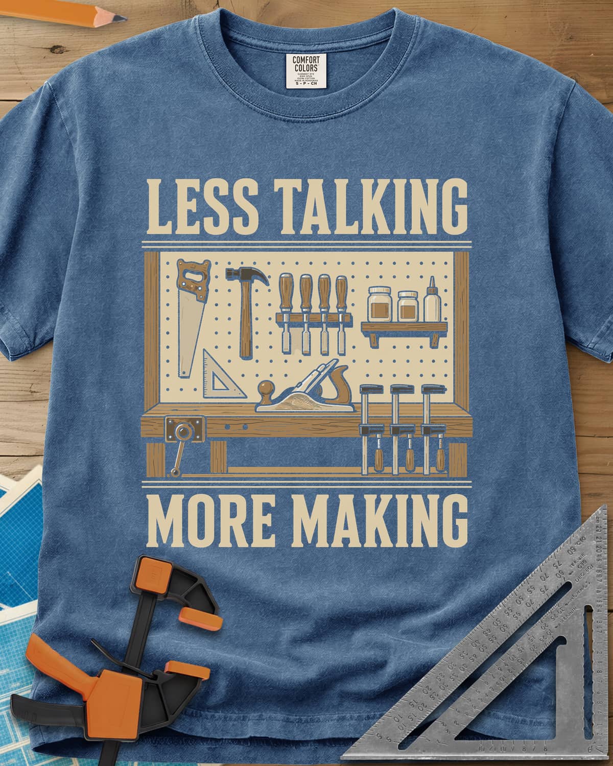 Less Talking More Making heavyweight tee in Blue Jean, a pegboard of hand tools over a workbench with a plane and clamps
