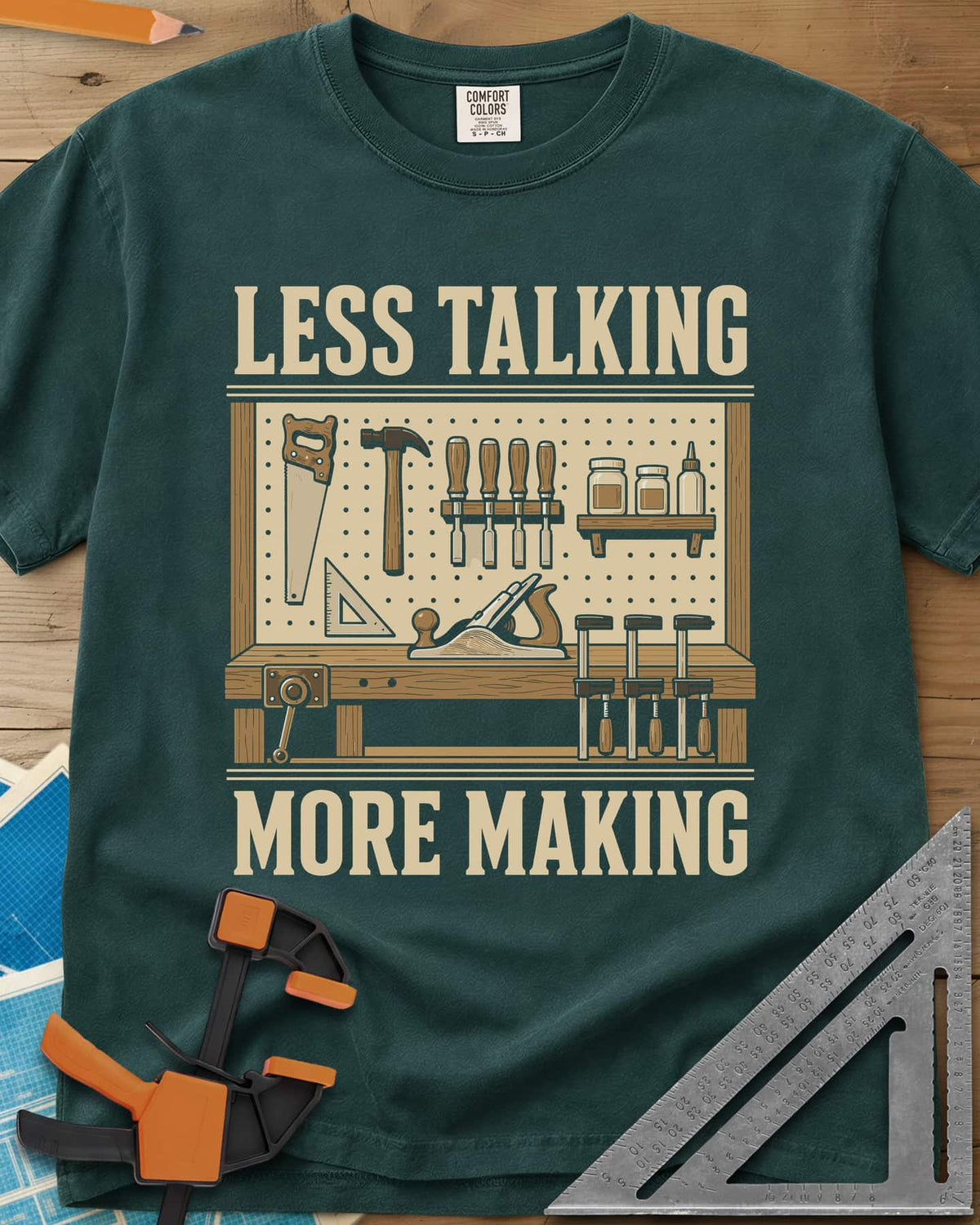 Less Talking More Making heavyweight tee in Blue Spruce, a pegboard of hand tools over a workbench with a plane and clamps