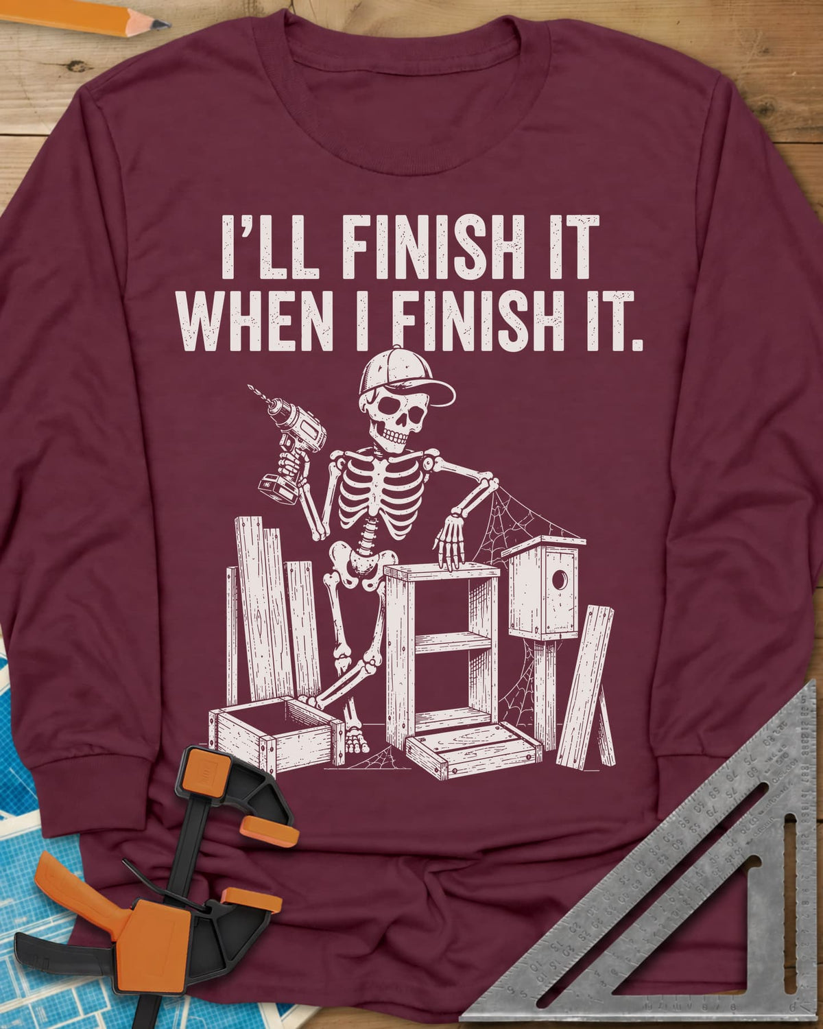 Misses Me At Work long sleeve shirt in Maroon, a skeleton in a ball cap with a drill among half-built projects