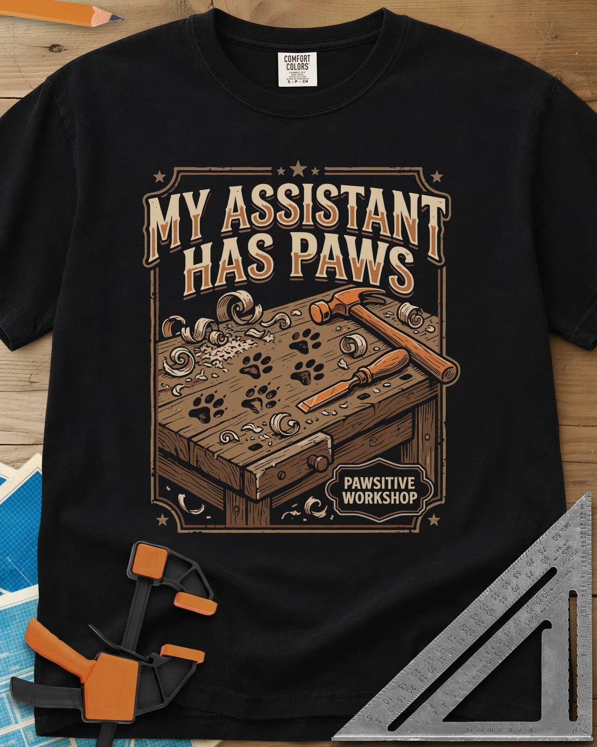 My Assistant Has Paws heavyweight tee in Black, workbench with paw prints, shavings, a hammer and a chisel