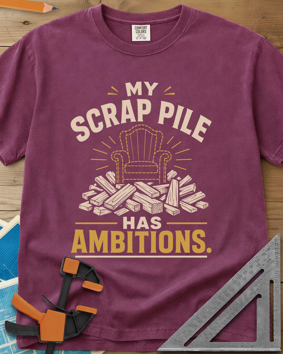 My Scrap Pile Has Ambitions heavyweight tee in Berry, a dotted gold armchair outline rising from a pile of wood offcuts