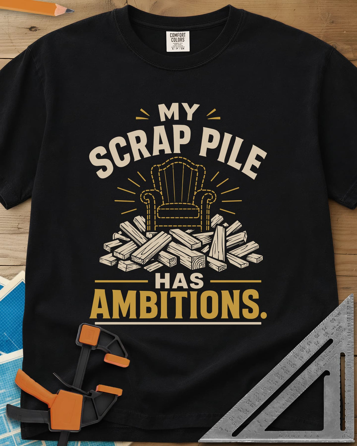 My Scrap Pile Has Ambitions heavyweight tee in Black, a dotted gold armchair outline rising from a pile of wood offcuts