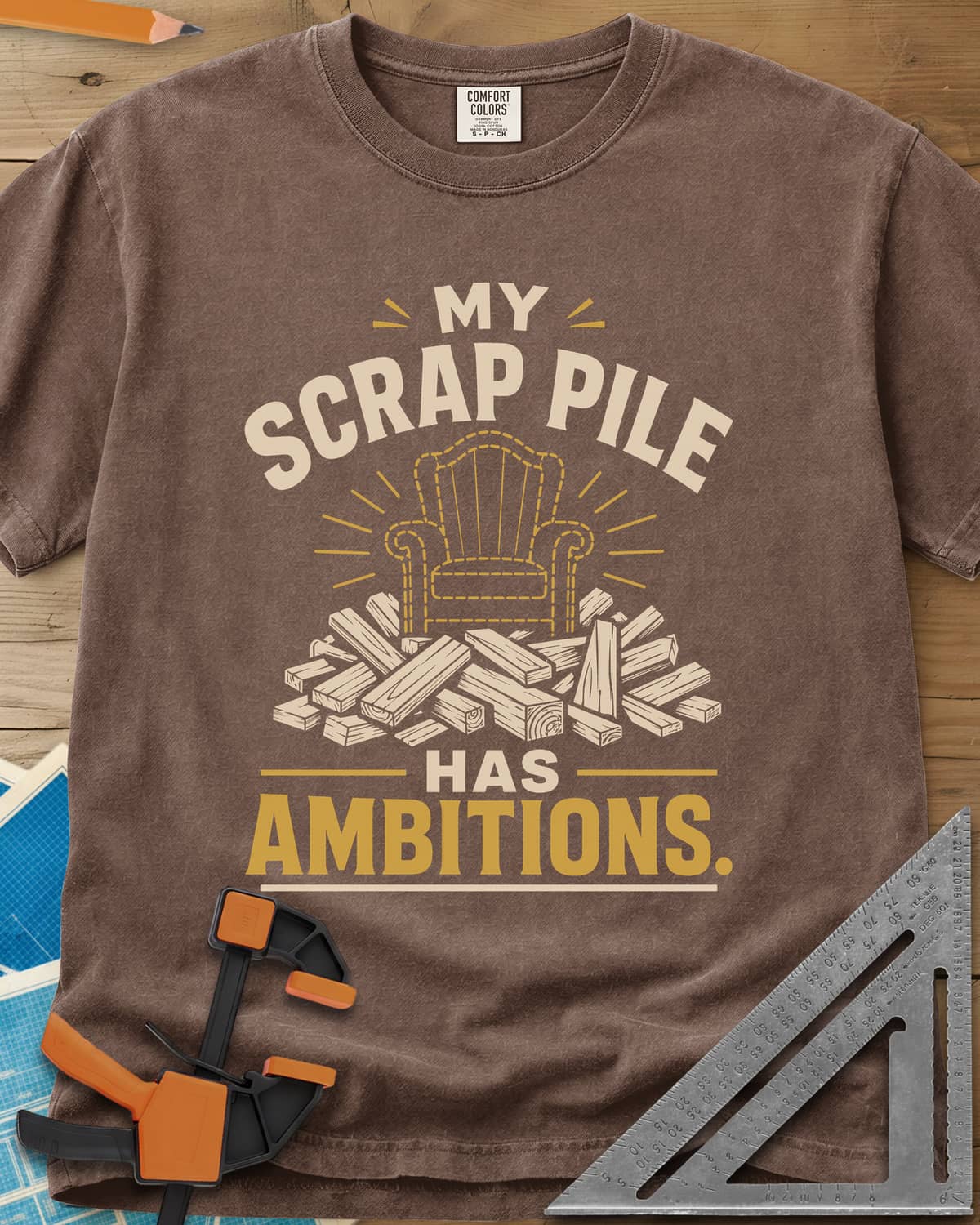 My Scrap Pile Has Ambitions heavyweight tee in Espresso, a dotted gold armchair outline rising from a pile of wood offcuts