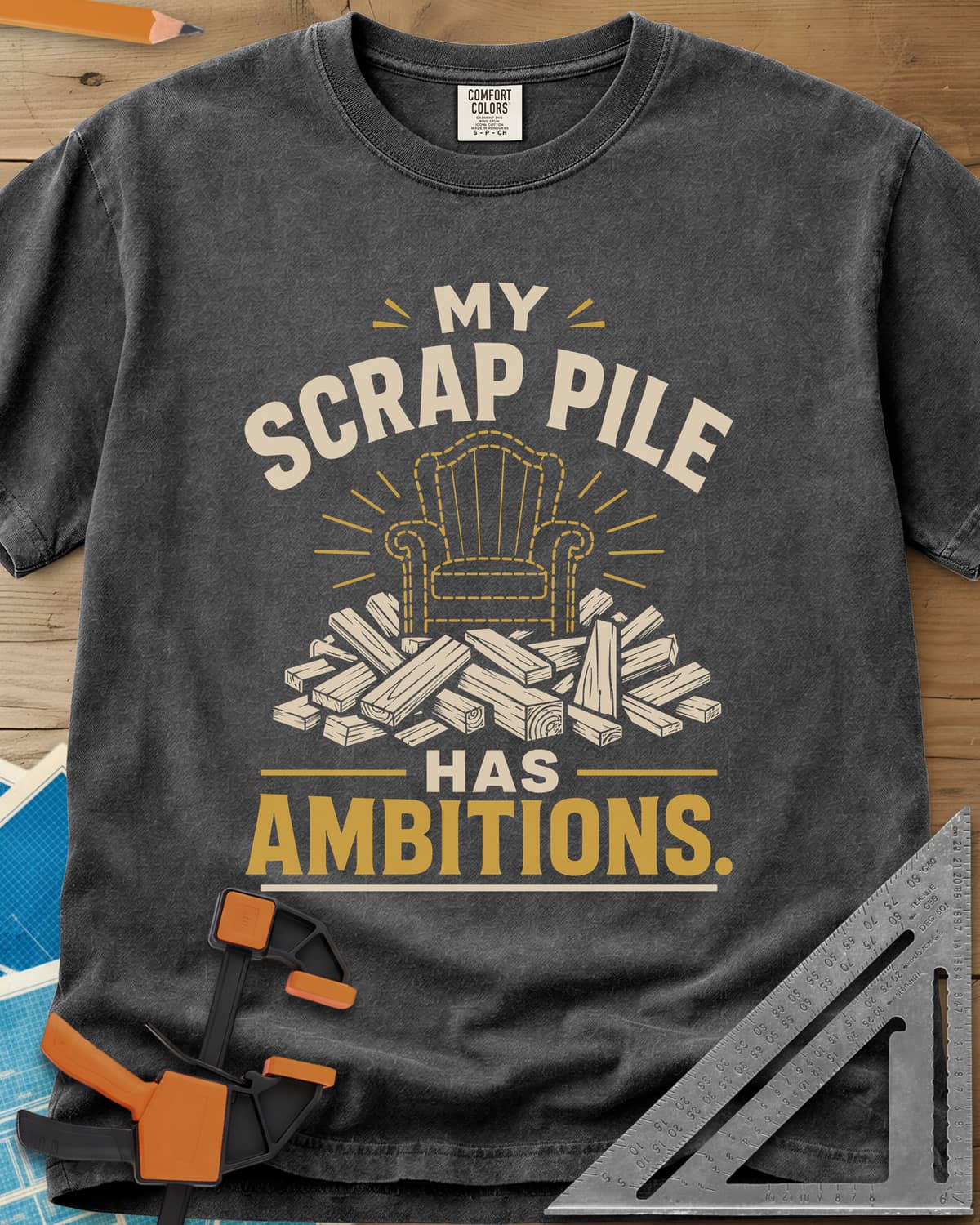 My Scrap Pile Has Ambitions heavyweight tee in Pepper, a dotted gold armchair outline rising from a pile of wood offcuts