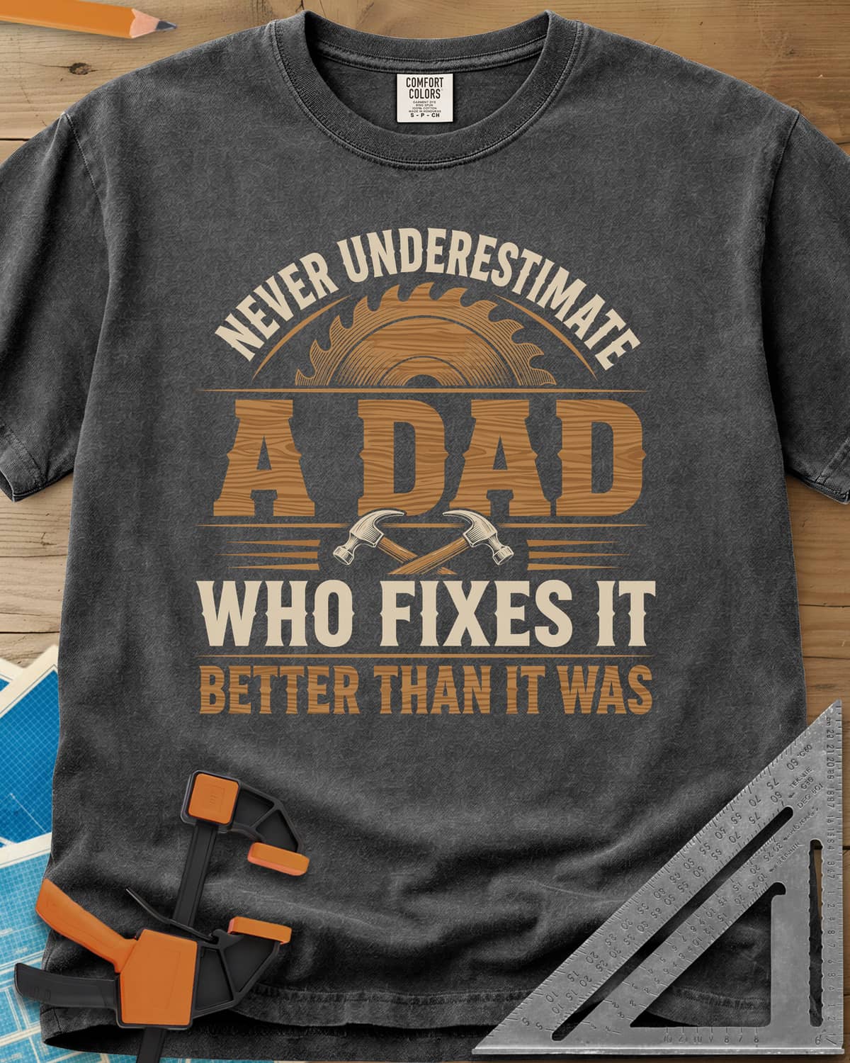 Never Underestimate A Dad Who Fixes It heavyweight tee in Pepper, saw blade and two crossed hammers around wood-grain