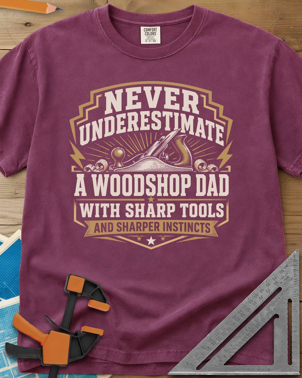 Never Underestimate A Woodshop Dad heavyweight tee in Berry, a shield badge with a hand plane, wood curls and sunburst rays