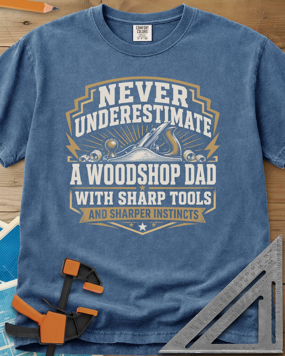 Never Underestimate A Woodshop Dad heavyweight tee in Blue Jean, a shield badge with a hand plane, wood curls and sunburst