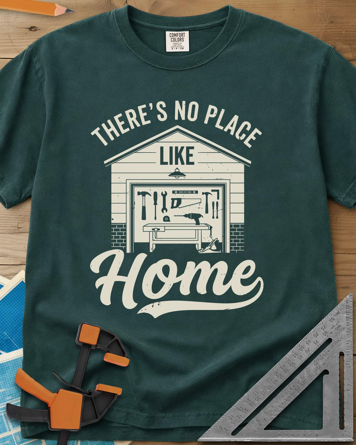 No Place Like Home heavyweight tee in Blue Spruce, a garage with its door up on a tool wall, above script Home