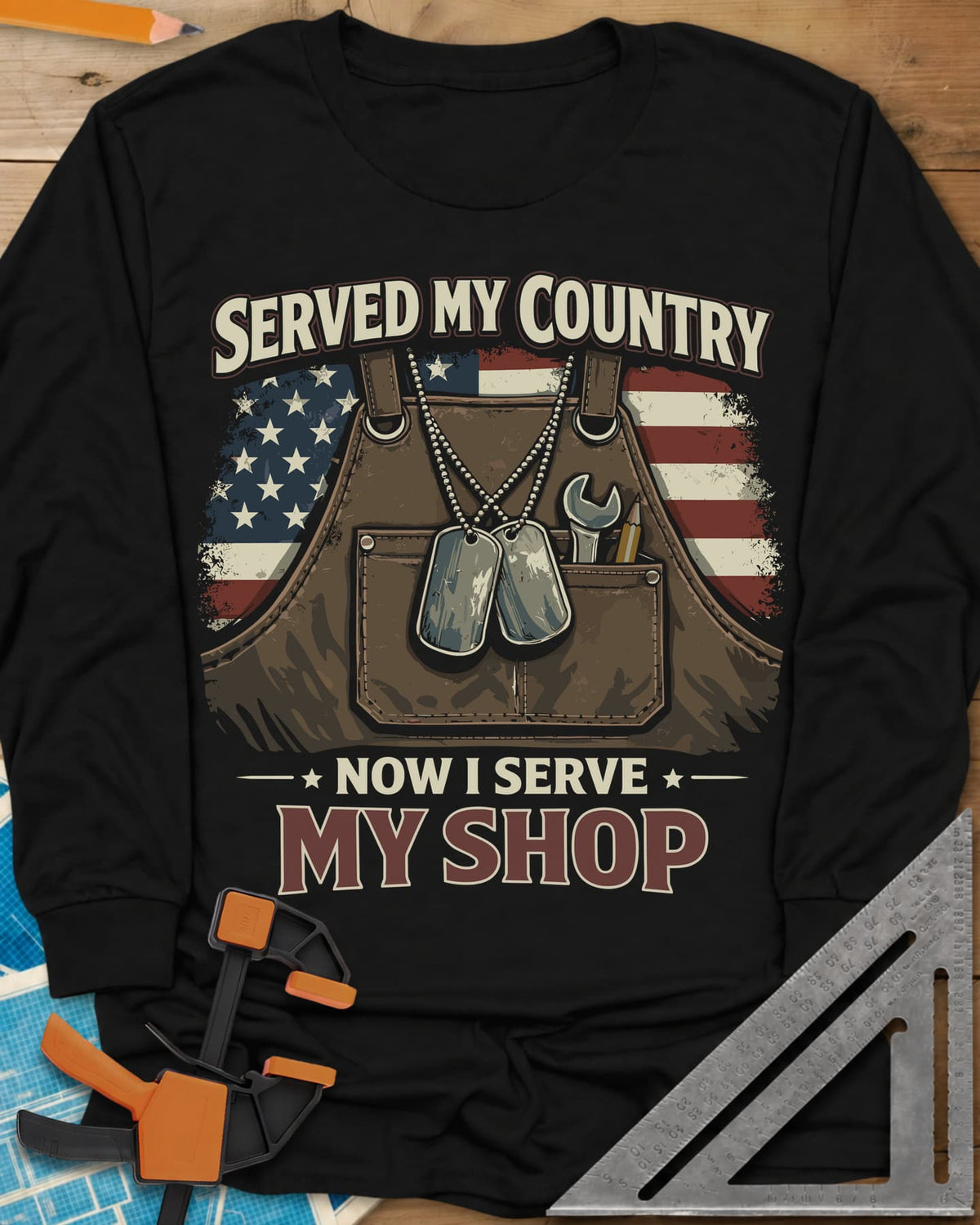 Now I Serve My Shop long sleeve shirt in Black, a shop apron with dog tags, a wrench and a pencil before a flag