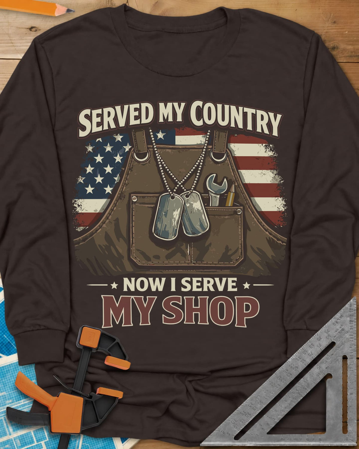 Now I Serve My Shop long sleeve shirt in Dark Chocolate, a shop apron with dog tags, a wrench and a pencil before a flag