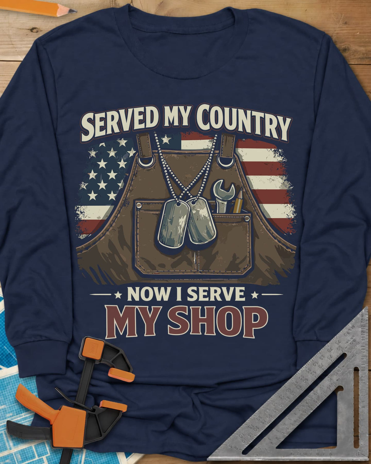 Now I Serve My Shop long sleeve shirt in Navy, a shop apron with dog tags, a wrench and a pencil before a flag