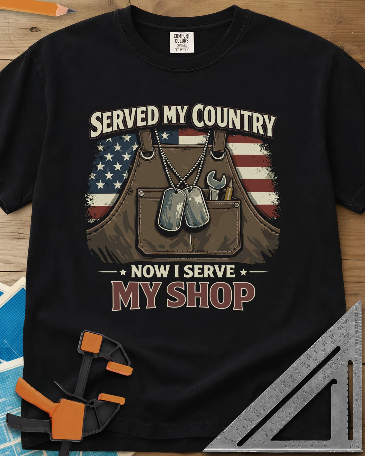 Now I Serve My Shop heavyweight tee in Black, a shop apron with dog tags, a wrench and a pencil before a flag