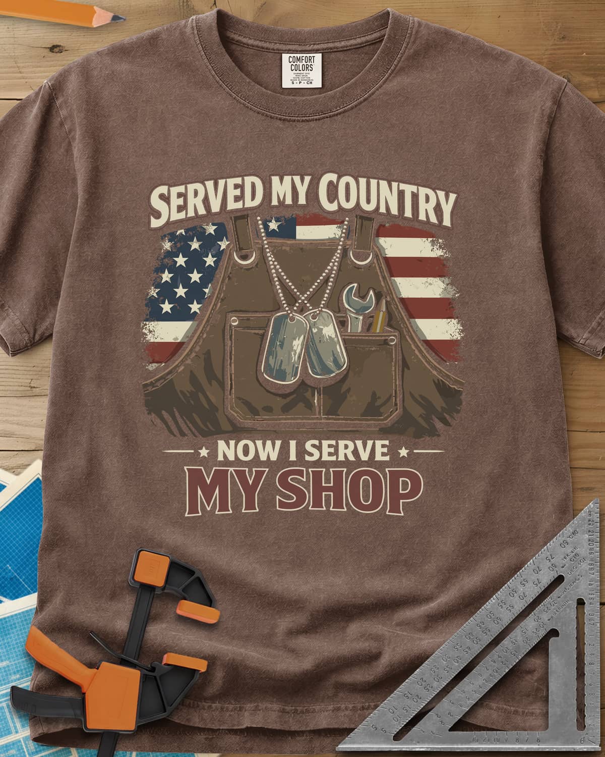 Now I Serve My Shop heavyweight tee in Espresso, a shop apron with dog tags, a wrench and a pencil before a flag
